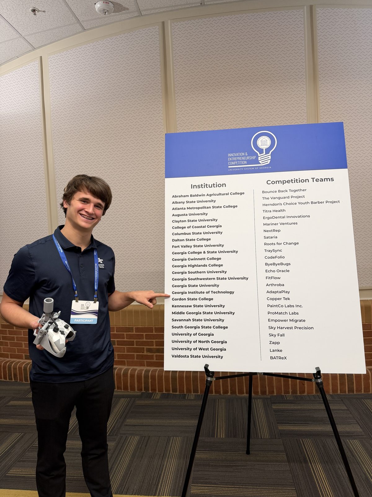 Ákos Vida holding the adaptive controller add-on and pointing to Georgia Tech on the board listing the competing institutions and teams.