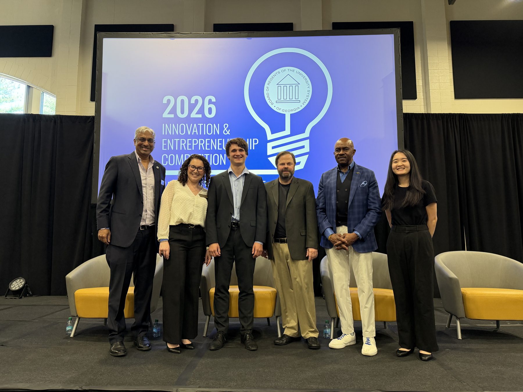 Ákos Vida standing with the judges on stage in front of the 2026 USG Innovation & Entrepreneurship Competition banner.
